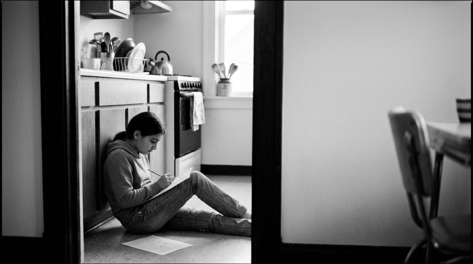 A child sitting on the kitchen floor against the cabinets, seen through a doorway, writing on a worksheet on her knees